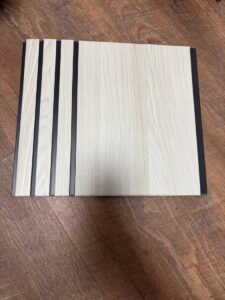 Light white wood-texture fluted wall panel with wide vertical sections and dark recessed grooves.